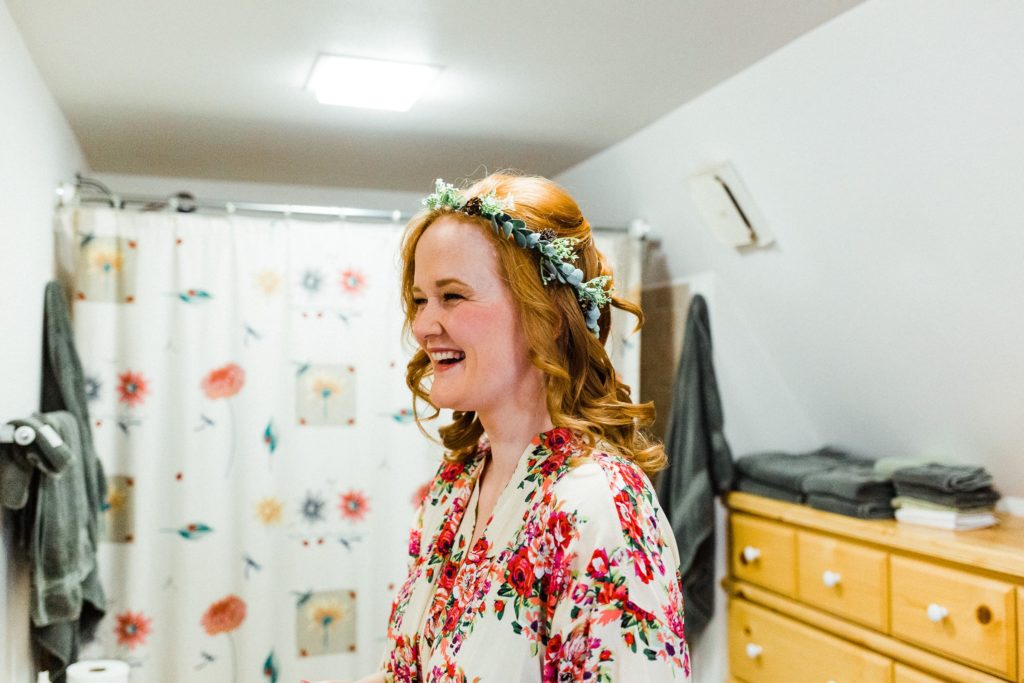 smiling bride wearing a flower crow is getting ready in a bathroom.