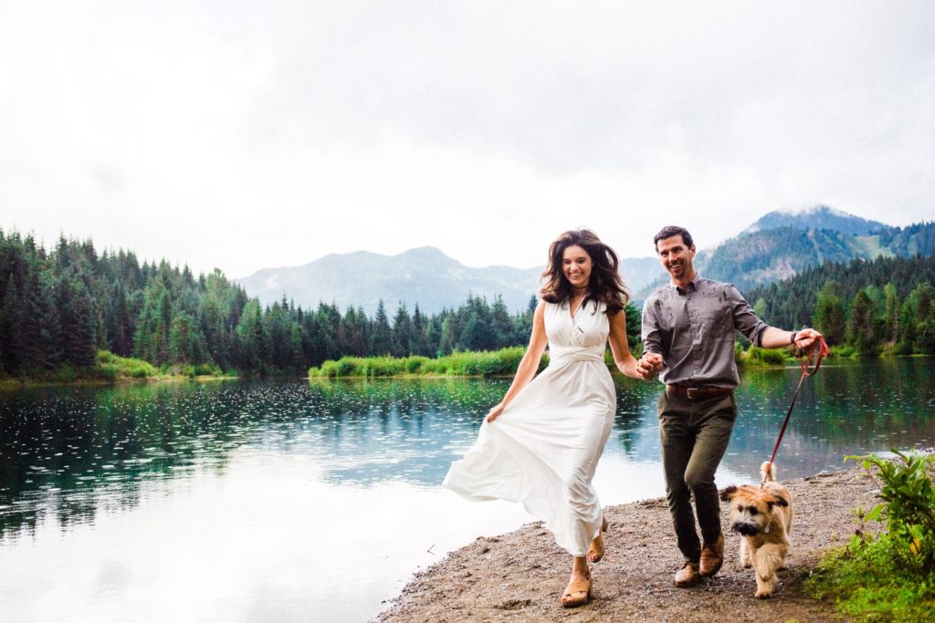couple running in wedding gear with dog by a mountain lake.