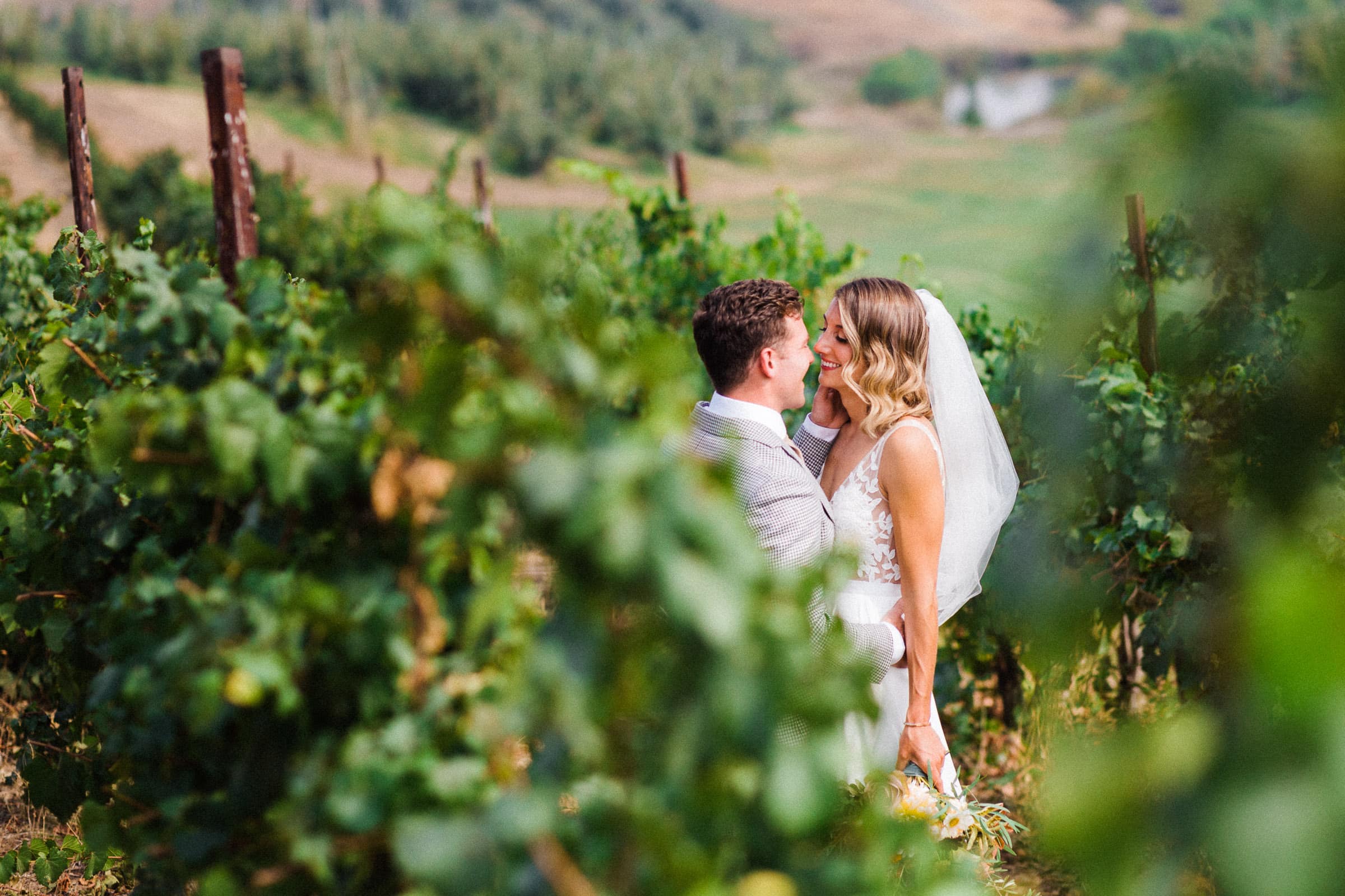 married couple smiling at each other between the vines in a vineyard