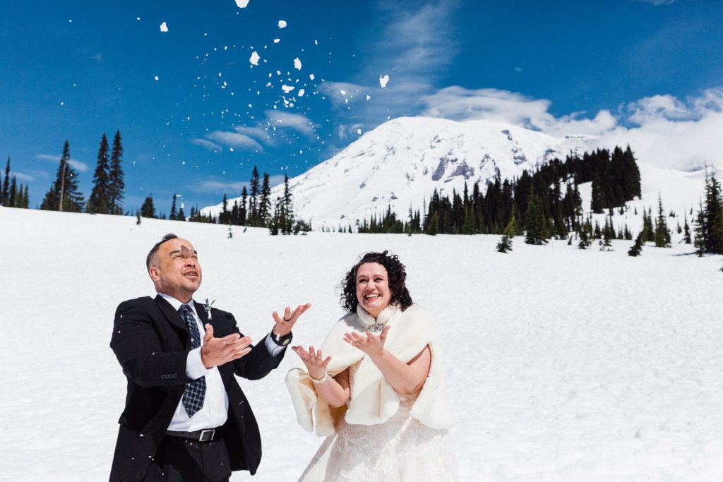couple in wedding attire throwing snow on a sunny day with mount rainier in the background