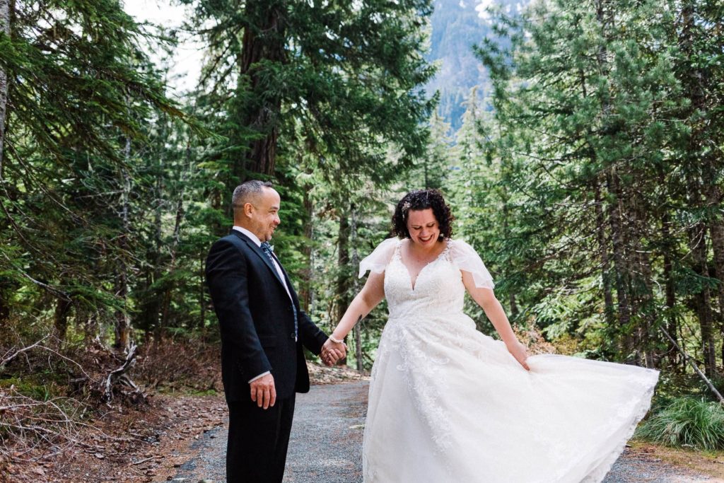 couple in wedding attire, laughing in a forest as she throws her dress